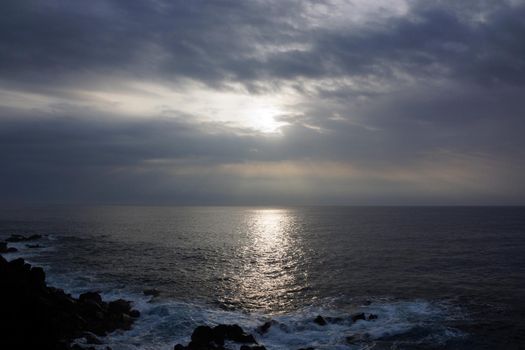 Sunrise over the ocean through the clouds with waves crashing along rocky shore on a voggy Big Island Hawaii day with coconut trees hanging over the water.                               