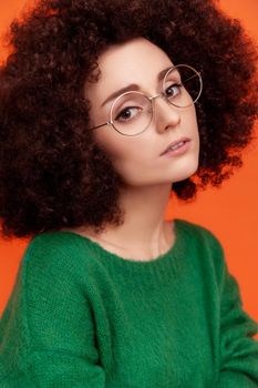 Portrait of pensive woman with perfect skin with Afro hairstyle wearing green casual style sweater, looking at camera, beauty and care concept. Indoor studio shot isolated on orange background.