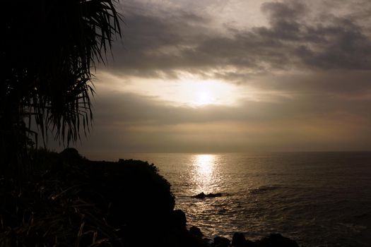 Sunrise over the ocean with waves crashing along rocky shore on a voggy Big Island Hawaii day with Lauhala trees silhouette along the shore.