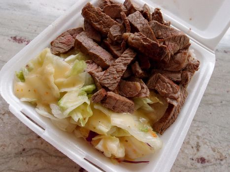 Large Steak and Salad Plate lunch in Styrofoam container on marble table with lettuce, purple cabbage, and Italian dressing.