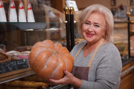 Happy senior female shop assistant laughing joyfully holding a pumpkin working at her store