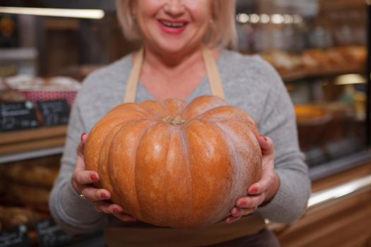 Cropped close up of a cheerful senior woman holding out a pumpkin to the camera, working at her groceries store
