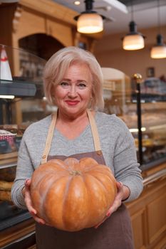 Vertical shot of a cheerful elderly shop assistant smiling joyfully, holding a pumpkin