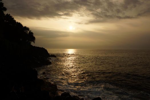 Sunrise over the ocean with waves crashing along rocky shore on a voggy Big Island Hawaii day with coconut trees lining the shore.                               