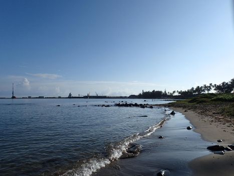 water along beach shore of Kahului Bay with coconut trees, and shipping cranes in the distance on West Maui on a beautiful morning day.