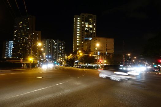 Cars race along Kapiolani Boulevard at Night with high-rises along street in Honolulu, Hawaii.