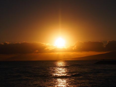 Dramatic Sunset over the clouds and reflecting on the Pacific ocean on the water with airplane flying off the coast of Oahu, Hawaii with Waianae Mountain range visible.