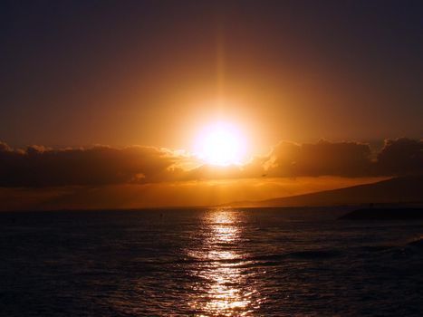 Dramatic Sunset over the clouds and reflecting on the Pacific ocean on the water with airplane flying off the coast of Oahu, Hawaii with Waianae Mountain range visible.