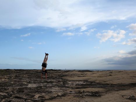 Man wearing a t-shirt, shorts, and slippers Handstands on shore rocks of Ko Olina on Oahu, Hawaii with dramatic clouds, sky, and pacific ocean in the distance. 