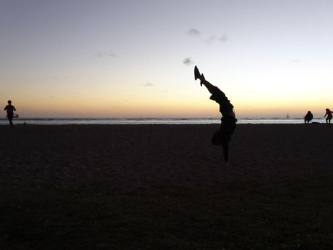 Man wearing t-shirt, shorts, and slippers Man Handstands on Ala Moana Beach at Dusk with boats in the ocean in the City of Honolulu on Oahu, Hawaii