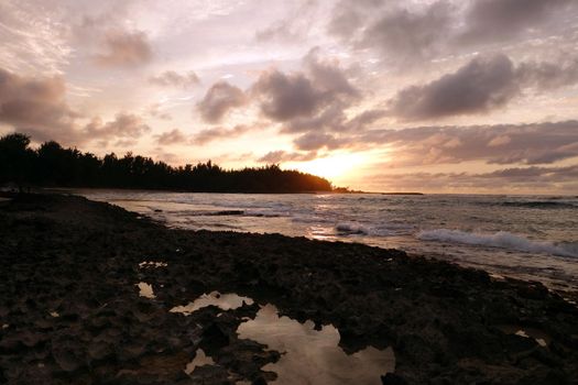 Sunset through the clouds and reflecting on the waves as they break on rocky shore with tide pools at Kawela Bay on the North Shore of Oahu. 