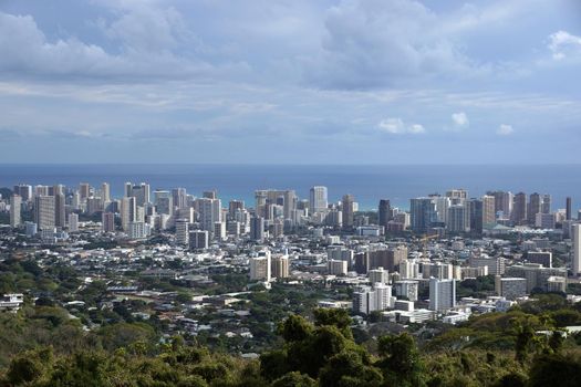 Honolulu cityscape, roads, buildings, skyscrapers, cranes, parks, and Pacific Ocean with clouds in the sky seen from up in the mountains on Oahu, Hawaii on a beautiful day. 