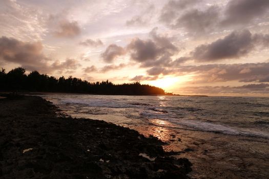 Sunset through the clouds and reflecting on the waves as they break on rocky shore at Kawela Bay on the North Shore of Oahu. 