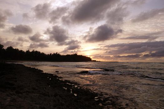 Sunset through the clouds and over the trees as it reflects on the watery waves at Kawela Bay rocky shore on the North Shore of Oahu.