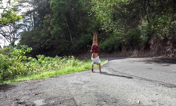 Man wearing a hat, t-shirt, shorts, and slippers Handstands at Tantalus Mountain road with lush vegetation above the City of Honolulu on Oahu, Hawaii