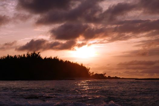 Sunset through the clouds and over the trees as it reflects on the watery waves at Kawela Bay on the North Shore of Oahu.