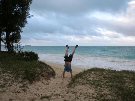 Man does handstand in the sand wearing t-shirt and shorts on the Waimanalo beach on Oahu with his legs open with a cloudy sky. 