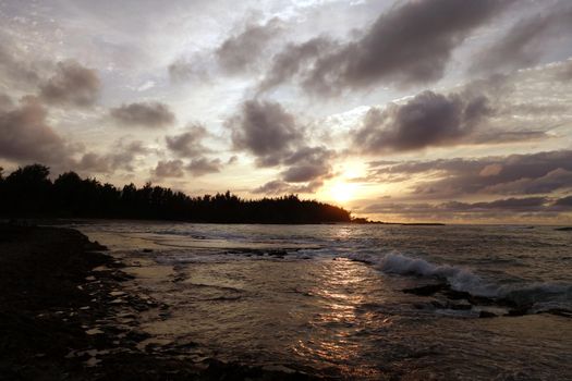 Sunset through the clouds and over the trees as it reflects on the watery waves at Kawela Bay rocky shore on the North Shore of Oahu.