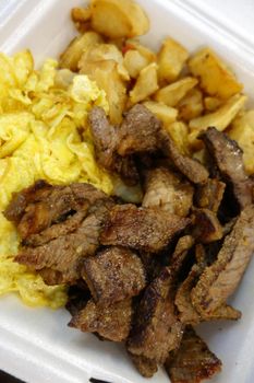 Kiawe grilled steak, scrambled eggs, country fried potatoes in a styrofoam plate on a tray on a table.