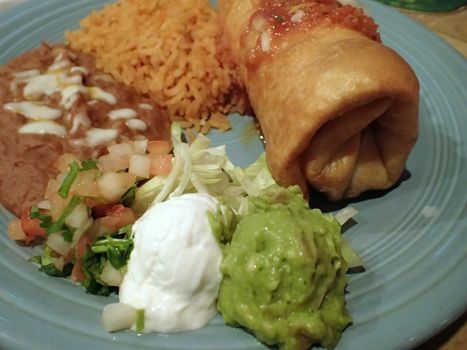 mexican chimichanga plate featuring spanish rice, refried beans, guacamole, pico de gallo, and sour cream on a blue plate.