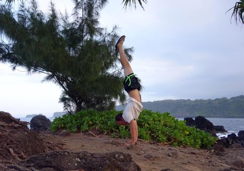 Man wearing a hat, t-shirt, pants, and slippers Handstands on Rocky shore with Kainalimu Bay in the background on Maui, Hawaii