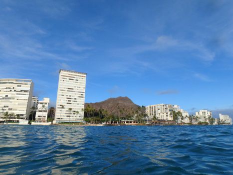 Hotel building, Outrigger Canoe Club, coconut trees, Condo buildings, clouds, and Diamond Head Crater in the distance on Oahu, Hawaii viewed from the water on a beautiful day. 