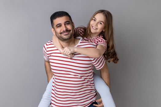 Portrait of positive optimistic father and daughter in striped T-shirts spending time together, dad holding his charming little kid on his back. Indoor studio shot isolated on gray background.