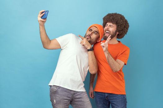 Portrait of two handsome smiling young adult hipster men pointing to camera and showing v sign while having video call or broadcasting livestream. Indoor studio shot isolated on blue background.