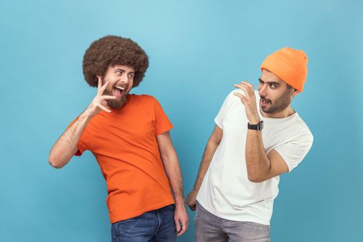 Portrait of two crazy young adult hipster men in T-shirts standing with raised claws, looking at each other with funny facial expression. Indoor studio shot isolated on blue background.