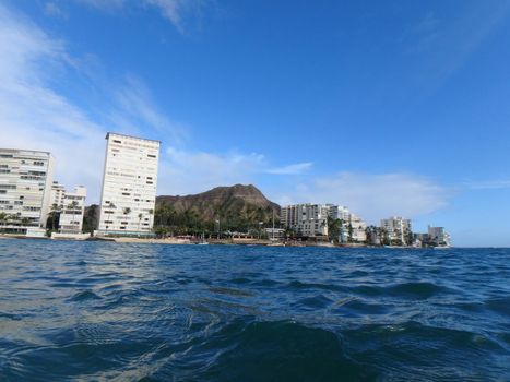 Hotel building, Outrigger Canoe Club, coconut trees, Condo buildings, clouds, and Diamond Head Crater in the distance on Oahu, Hawaii viewed from the water on a beautiful day. 