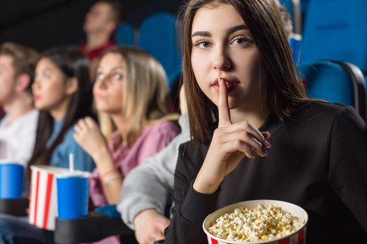 No speaking. Portrait of a beautiful young woman holding popcorn bucket making shushing gesture to the camera at the cinema