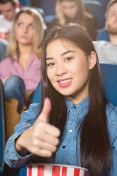 Movie approved. Young happy asian woman holding popcorn bucket showing thumbs up smiling cheerfully at the cinema