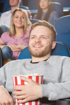 Cinema lover. Vertical shot of a handsome bearded young man holding popcorn bucket laughing watching a movie at the local cinema