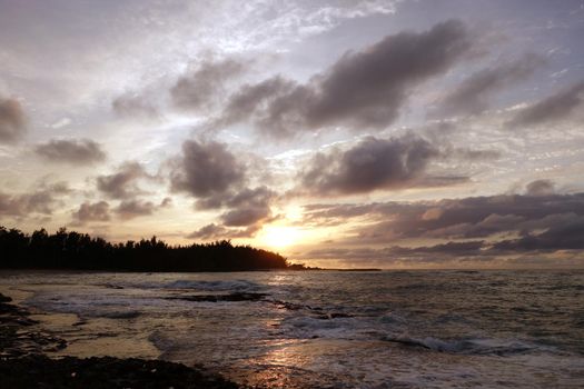 Sunset through the clouds and over the trees as it reflects on the watery waves at Kawela Bay rocky shore on the North Shore of Oahu.