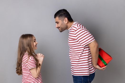 Portrait of happy festive father and daughter in striped T-shirts, man hiding gift box behind his back, little girl waiting for her present. Indoor studio shot isolated on gray background.