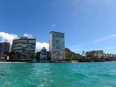 Hotel building, Outrigger Canoe Club, coconut trees, Condo buildings, clouds, and Diamond Head Crater in the distance on Oahu, Hawaii viewed from the water on a beautiful day. 