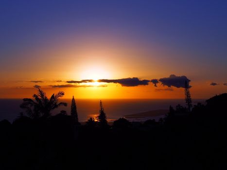 Sunset through the clouds over the pacific ocean with light reflecting on water past tropical silhouette of trees on Oahu, Hawaii.  February 2016.