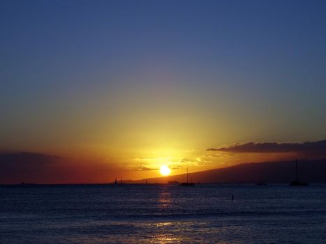 Dramatic lighting as Sunsets behind Waianae mountains with light reflecting on ocean and illuminating the sky with boats sailing on the water off Waikiki on Oahu, Hawaii. August 2015.