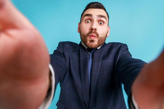 Funny bearded businessman wearing official style suit, taking selfie, looking at camera POV, point of view of photo, sending air kiss. Indoor studio shot isolated on blue background.