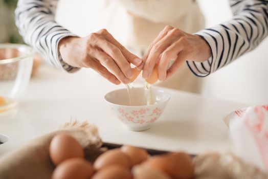 Horizontal shot of serious male wearing beige apron breaking egg into metal bowl