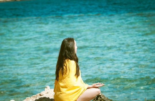 young woman in yellow bright scarf meditating at rocks at the sea