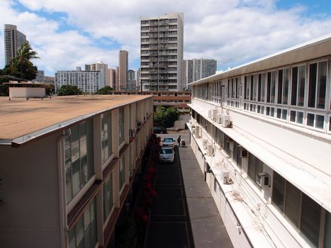 Alley between buildings with cars and Homeless person with shopping cart.  Highrises and surrounding Honolulu cityscape visible.