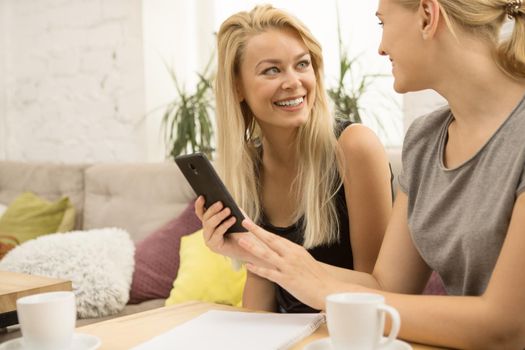 Beautiful young woman smiling at her friend cheerfully discussing something she is showing on a smart phone friendship gossip communication technology online internet social media girls lifestyle coffee shop