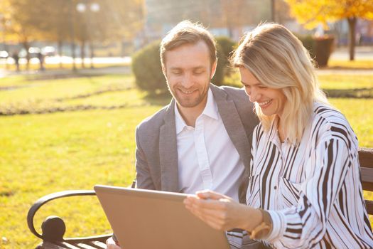 Two cheerful business colleagues enjoying working outdoors on a warm autumn day, using laptop together. Entrepreneurship, startup concept