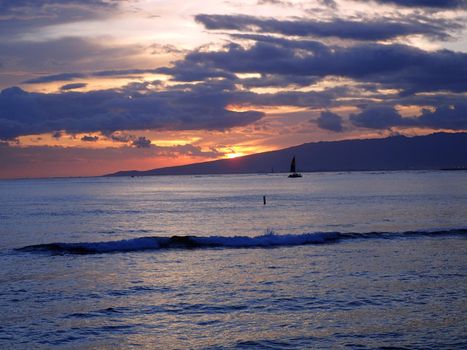 Dramatic Sunset dropping behind the Waianae Mountain range with clouds, waves and boats on the Pacific ocean off the coast of Oahu, Hawaii.