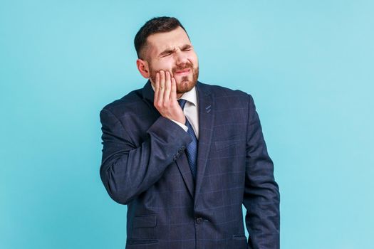 Unhappy bearded man wearing official style suit feeling toothache, touching sore cheek, suffering from cavities, cracked teeth, gum recession. Indoor studio shot isolated on blue background.