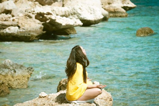 young woman in yellow bright scarf meditating at rocks at the sea