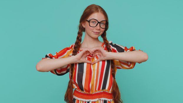Girl in love. Smiling redhead girl in dress makes heart gesture demonstrates love sign expresses good feelings and sympathy. Young one ginger teenager child isolated alone on blue studio background