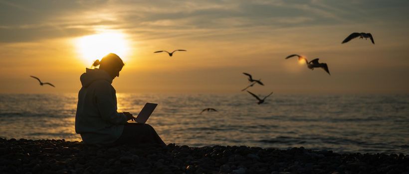 Caucasian woman typing on a laptop on the seashore at sunset. Freelance work. Wide screen