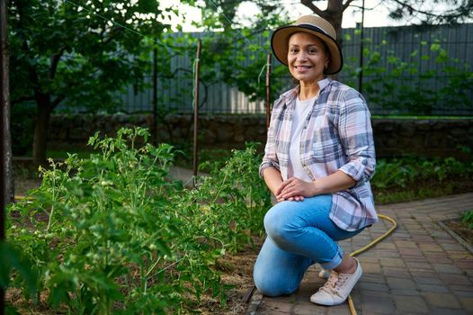 Attractive successful woman, owner of an ecological farm, sits next to tomato seedlings and smiles sweetly, looking at the camera. Beautiful prosperous young female farmer enjoying gardening in spring